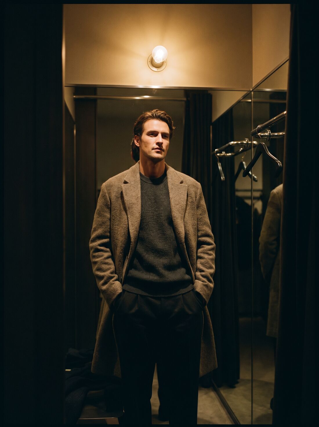 A man in a wool overcoat and fine knit in a dim fitting room